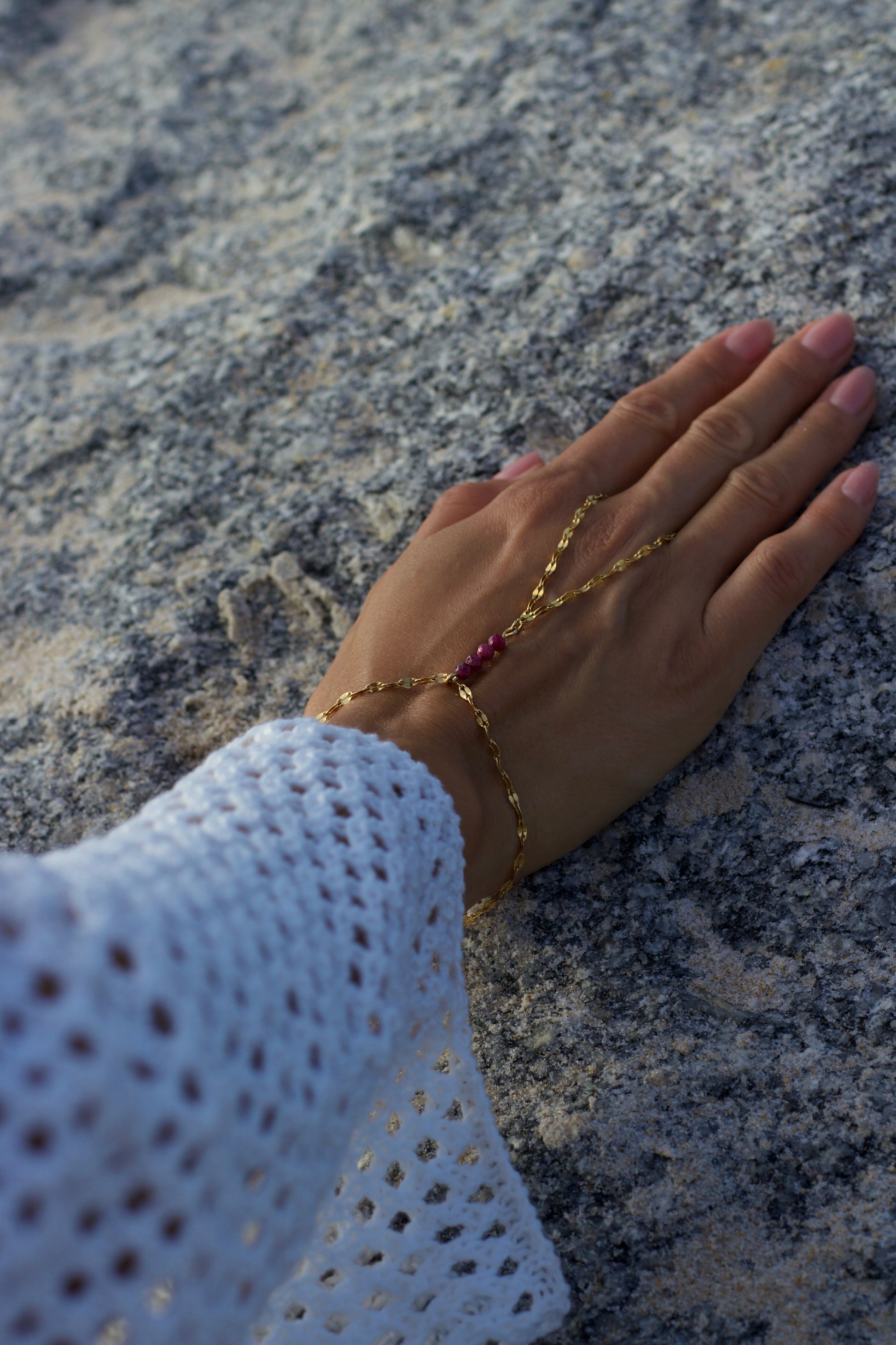 Hand wearing a gold bracelet with a red gemstone on a rocky surface