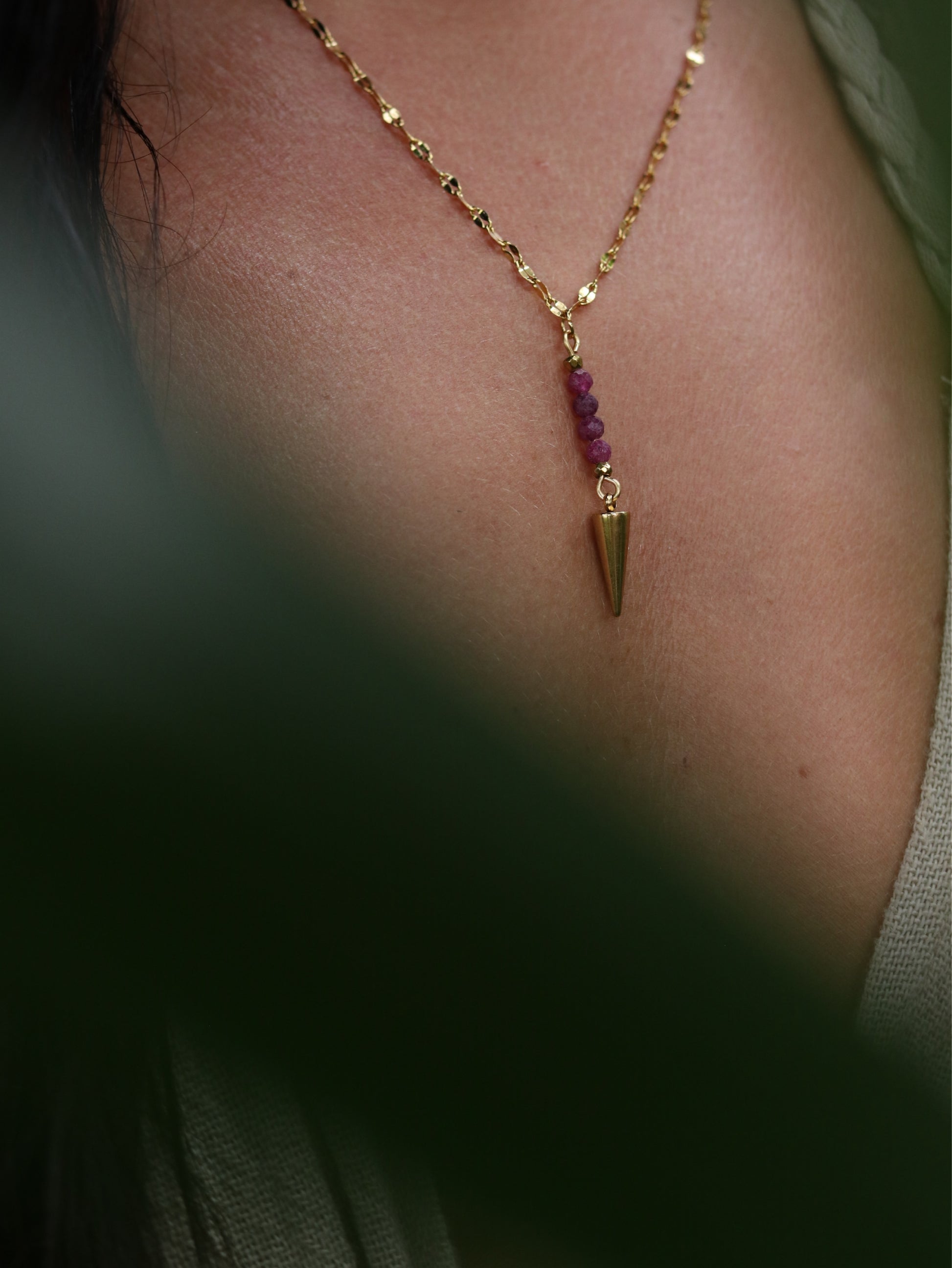 Gold necklace with a pendulum pendant and small ruby beads on a person's neck.
