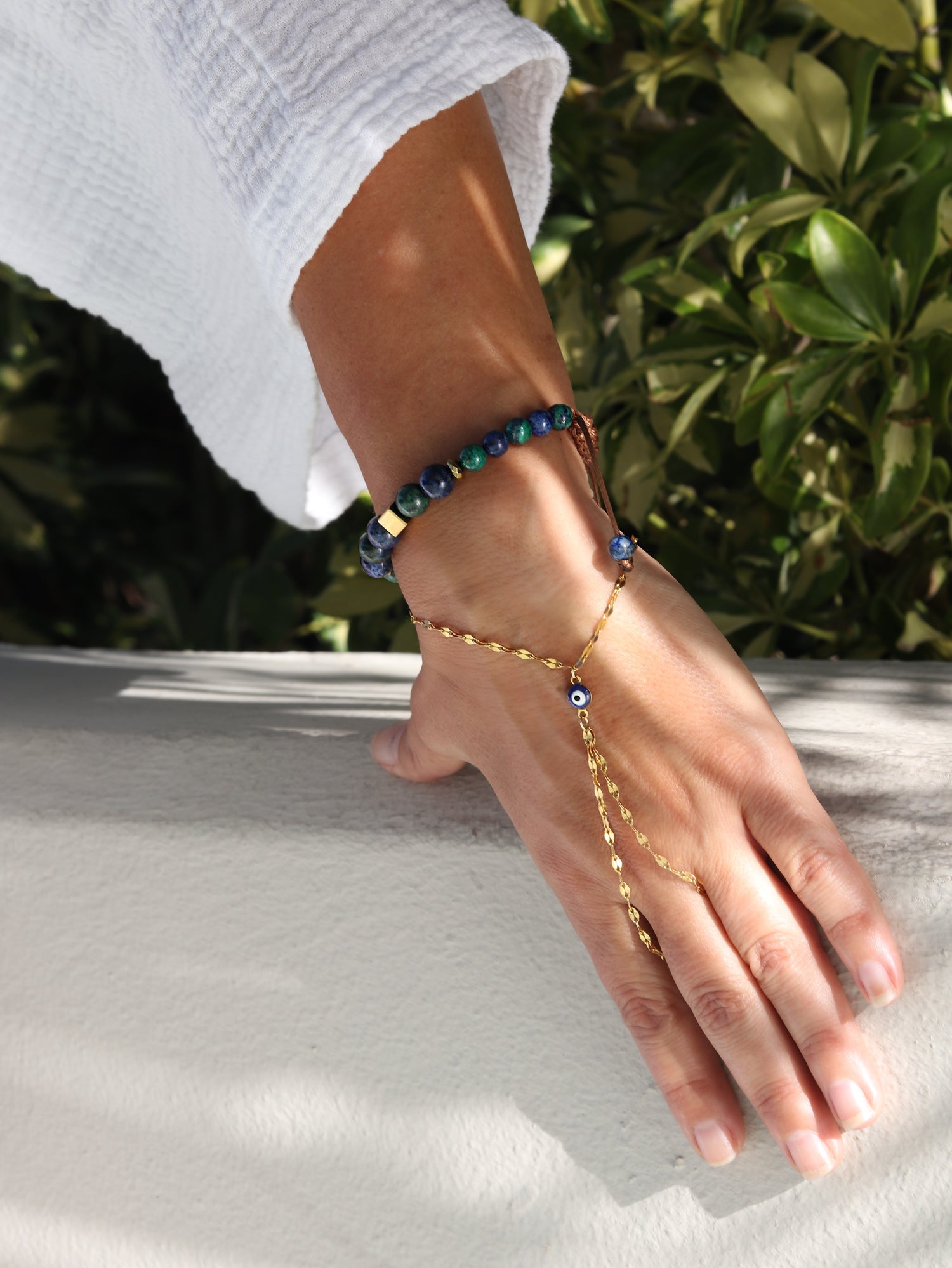 close up hand wearing beaded gemstone bracelet, and evil eye charm hand chain bracelets on a white surface with greenery in the background